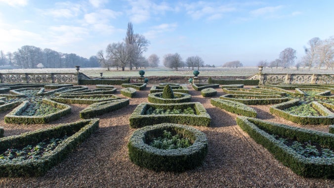 A view across the small box hedged parterre covered in a hard frost, over looking the river Avon, with wintry outlines of trees in the far distance against a cloud studded, blue sky.